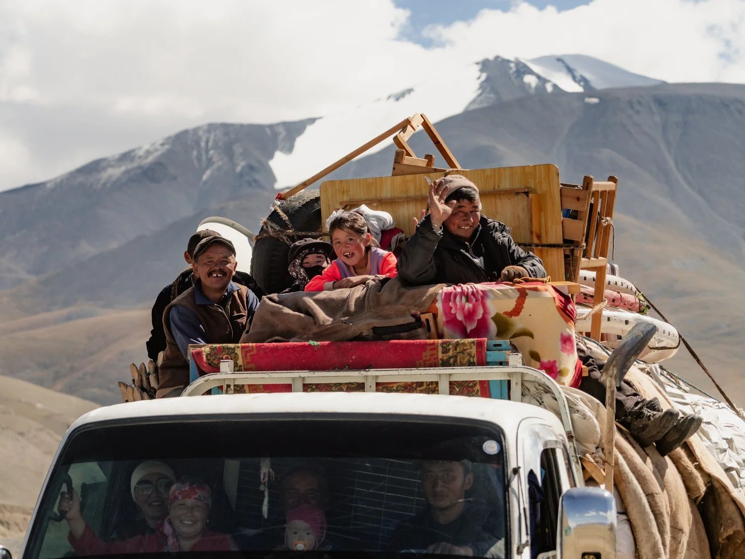 A nomad family waving from atop a loaded truck beneath snow-capped mountains in Mongolia